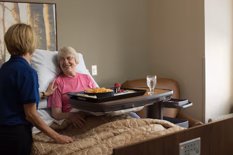 An elderly woman sits up in bed smiling at a caregiver while a meal tray and glass rest on an overbed table.