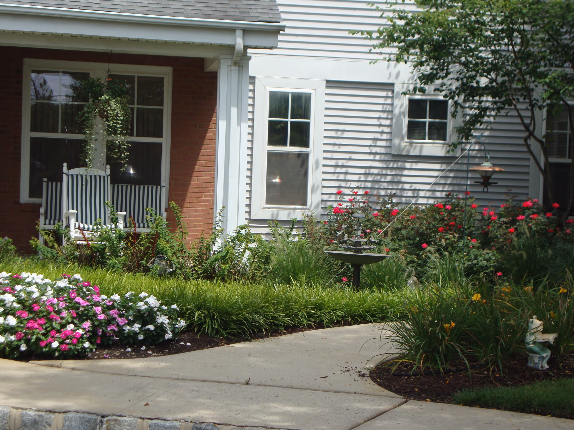 A garden area outside a building with a concrete walkway, colorful flower beds, green shrubs, a birdbath, and a white rocking chair with blue and white striped cushions on a porch. The building has white siding and brick walls with several windows.