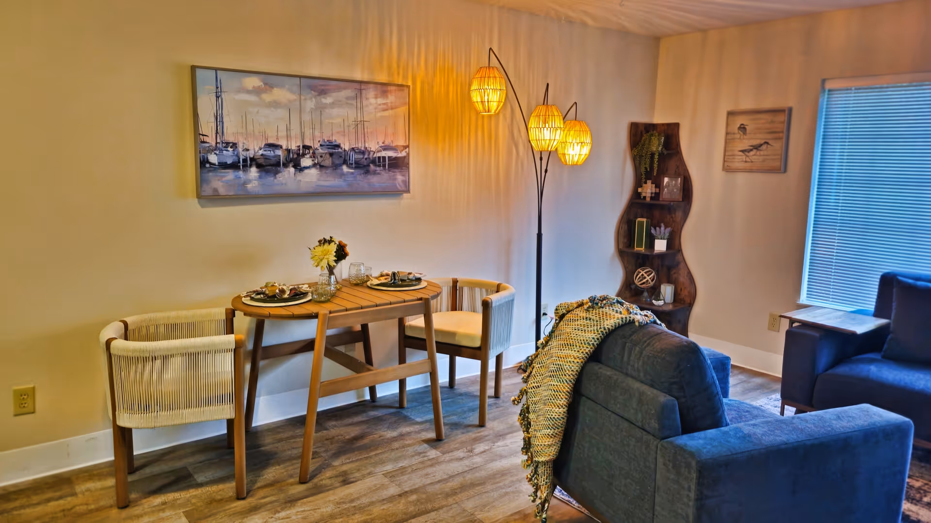 Cozy interior showing a small wooden dining table with two woven chairs, a blue sofa and armchair, a tall floor lamp, and wall art.