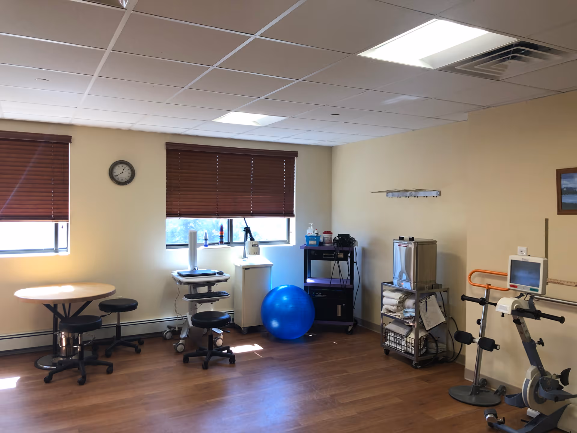 A rehabilitation room with wooden floors and beige walls, featuring exercise equipment including a stationary bike, a blue exercise ball, and various medical devices. There are two windows with brown blinds partially closed, a round table with stools, a clock on the wall, and shelves holding towels and supplies.