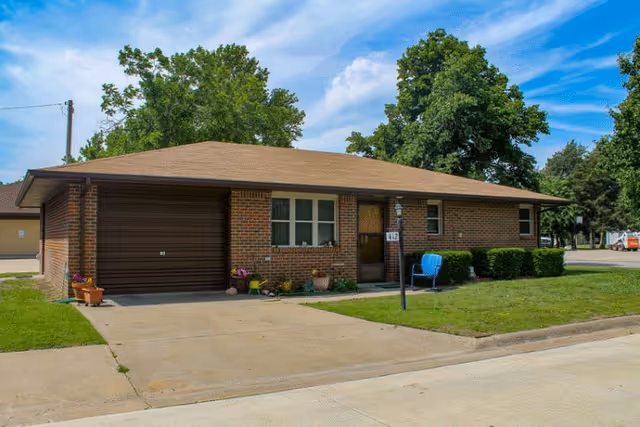 Single-story brick house with a brown roof and attached garage. There is a driveway leading up to the garage and a small lawn with trimmed bushes and a blue chair near the front door. Trees and a partly cloudy sky are visible in the background.