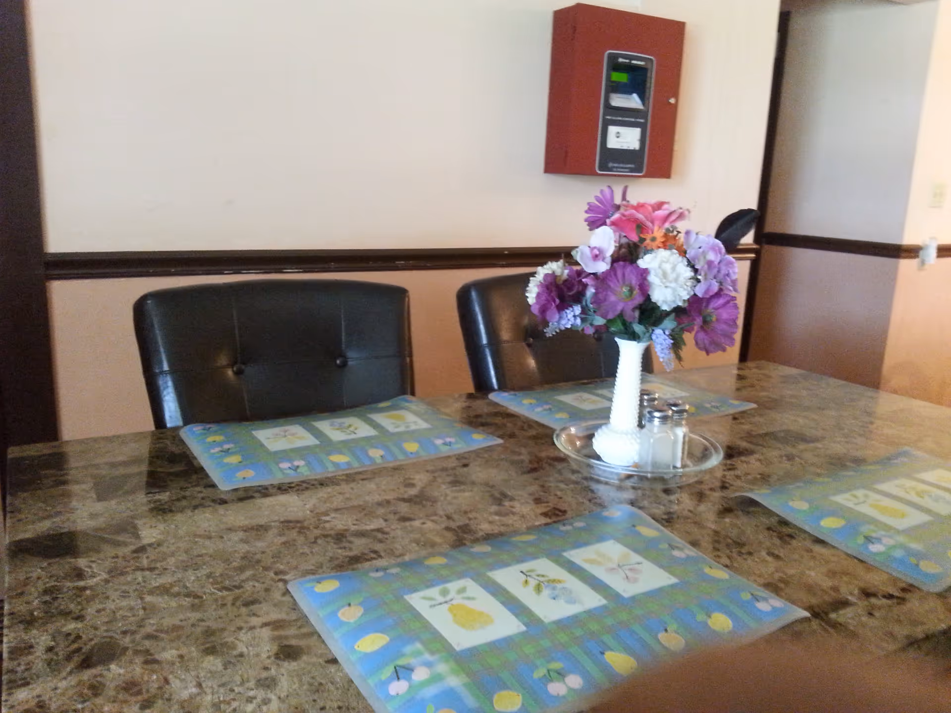 A dining table with a marble-like surface set with four colorful placemats featuring fruit designs. In the center of the table is a white vase with a bouquet of purple, pink, and white flowers, along with salt and pepper shakers on a clear tray. Two black cushioned chairs are visible, and a red wall-mounted device is on the beige wall in the background.