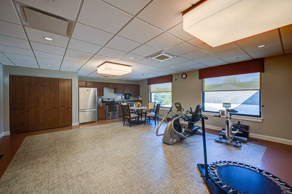 A spacious room with exercise equipment including a stationary bike, elliptical machine, and a mini trampoline. The room also features a small dining table with four chairs, a kitchen area with stainless steel refrigerator, stove, and cabinets. Large windows with red valances allow natural light to enter the room.
