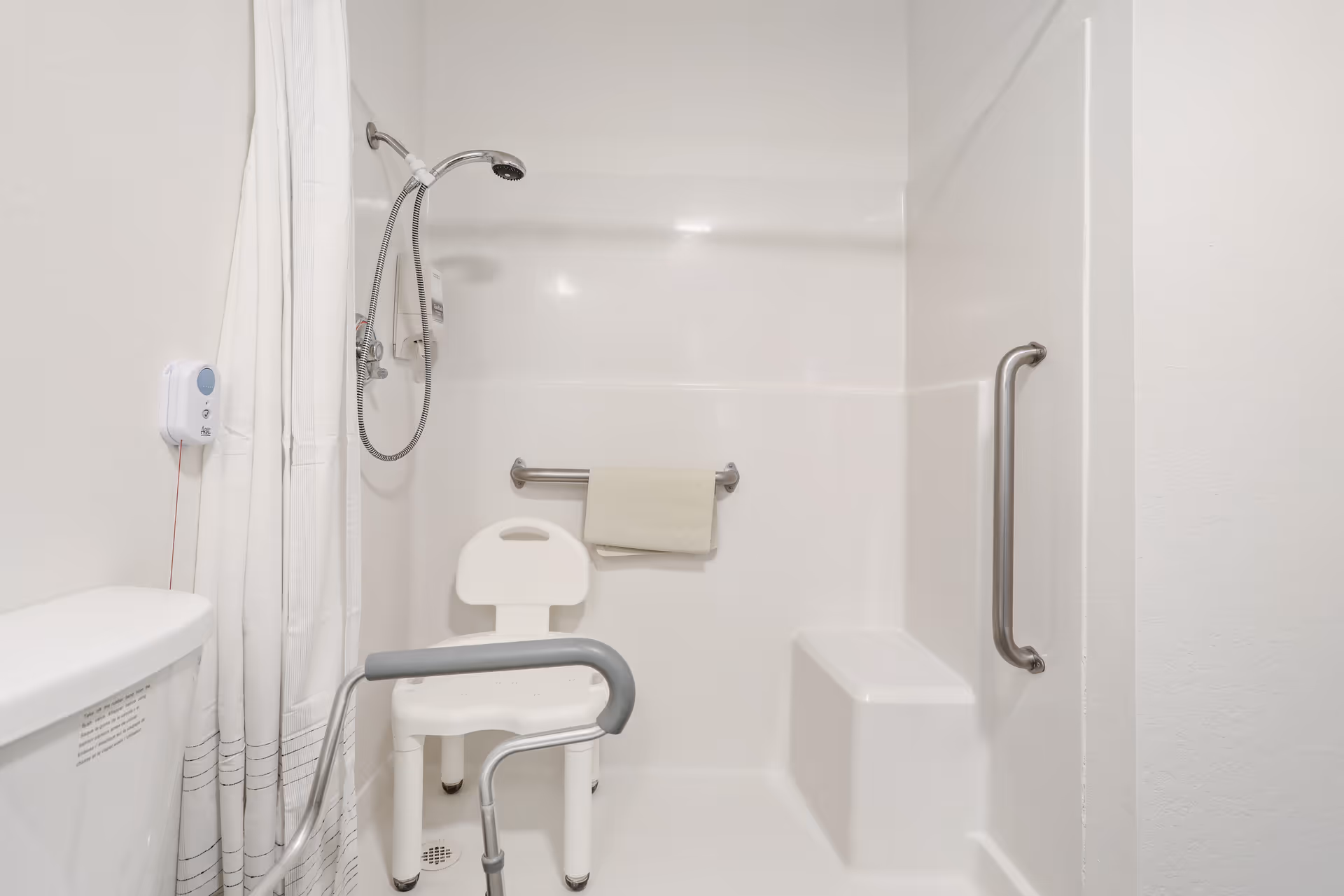 A white accessible shower area with a handheld showerhead, a white plastic shower chair, grab bars on the wall, a towel hanging on one grab bar, and a white shower curtain partially drawn. The shower area also includes a built-in seat and a toilet partially visible on the left side.