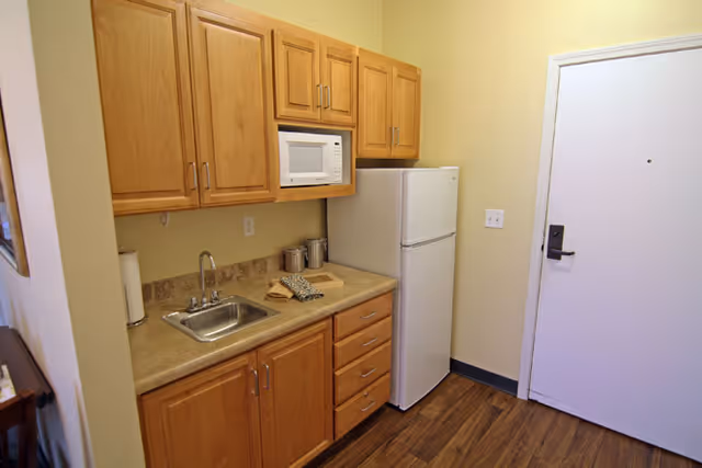 A small kitchen area with wooden cabinets, a countertop with a sink, a microwave mounted above the counter, and a white refrigerator next to a closed white door. The floor has wood-style flooring and the walls are painted light yellow.