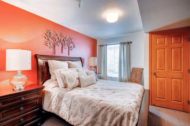 A cozy bedroom with a wooden bed frame and a neatly made bed featuring beige and white patterned bedding. There are two matching bedside tables with lamps on each side of the bed. The wall behind the bed is painted orange and decorated with a metal tree wall art. A window with light curtains allows natural light into the room, and a wooden door is visible on the right side.