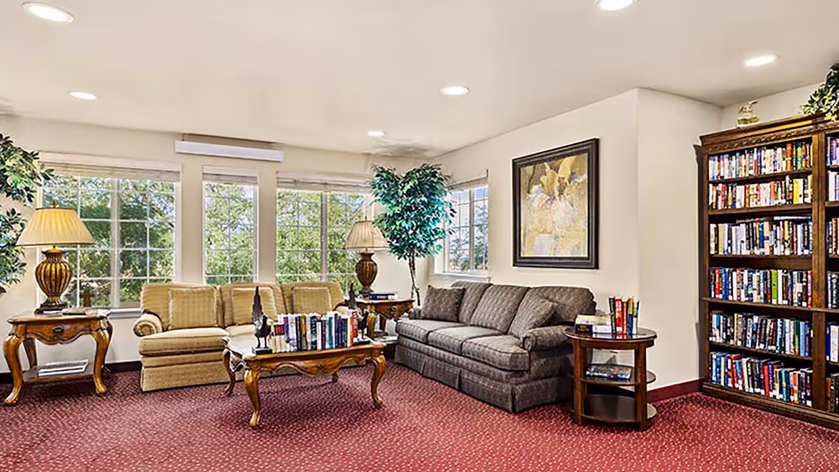 A cozy living room area with two sofas, a coffee table filled with books, two side tables with lamps, a large bookshelf filled with books, a framed painting on the wall, and large windows letting in natural light. The room has a red carpet and some indoor plants.