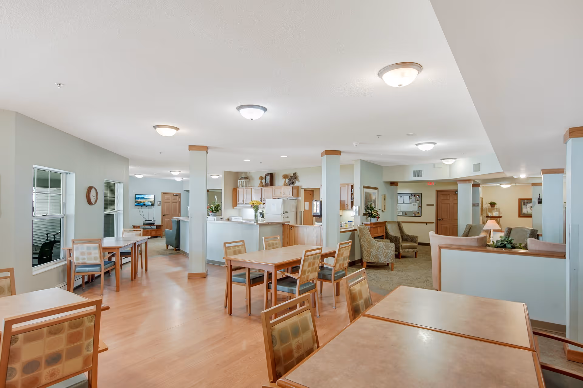 A spacious, well-lit common area in an assisted living facility featuring multiple wooden tables and chairs arranged for dining or socializing. The room has light-colored walls, wood flooring, and several ceiling lights. In the background, there is a kitchen area with wooden cabinets, a refrigerator, and a counter with flowers. There are also comfortable armchairs and a small seating area near the back of the room.