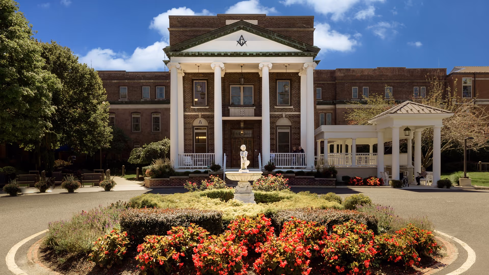 Front view of a brick building with large white columns, a circular flowerbed and fountain in the driveway.