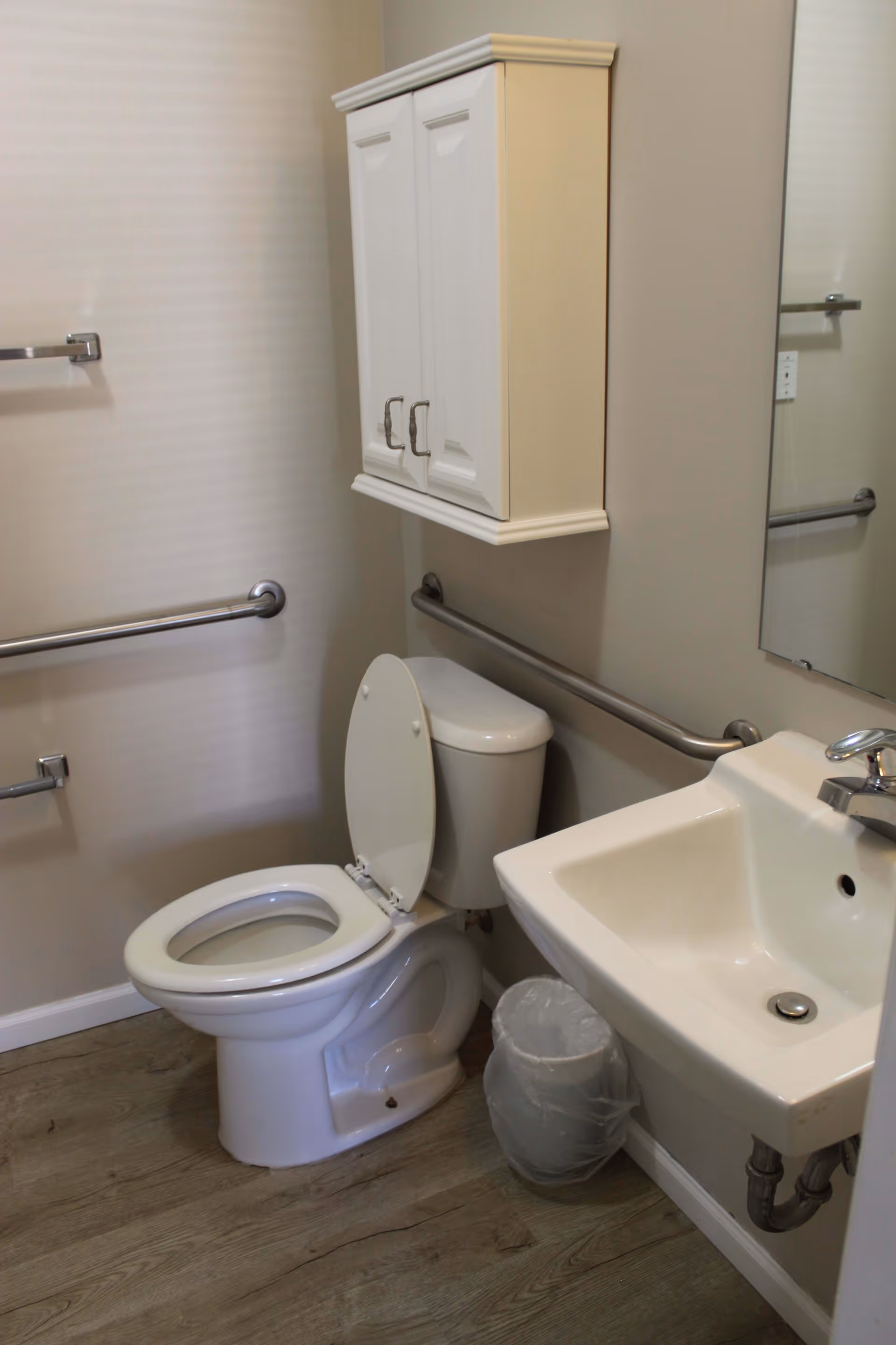 A small bathroom with a white toilet with the lid open, a white sink with a silver faucet, a wall-mounted white cabinet with two doors above the toilet, a trash can with a plastic liner, and metal grab bars on the walls for accessibility. The floor has a wood-like finish.
