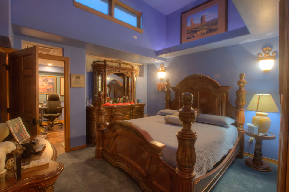 A bedroom with ornate wooden furniture including a large bed with carved posts, a matching dresser with a mirror, and a side table with a lamp. The walls are painted blue and decorated with framed pictures. An open door reveals an office area with a chair and desk.