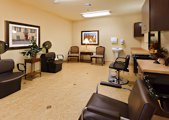 Interior view of a salon area in a senior living facility with multiple brown salon chairs, a small sink, a counter with cabinets, two framed paintings on the walls, and a small table with two chairs and a lamp in the background.