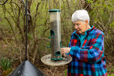 An older adult in a colorful plaid jacket refilling a hanging bird feeder in a wooded yard.