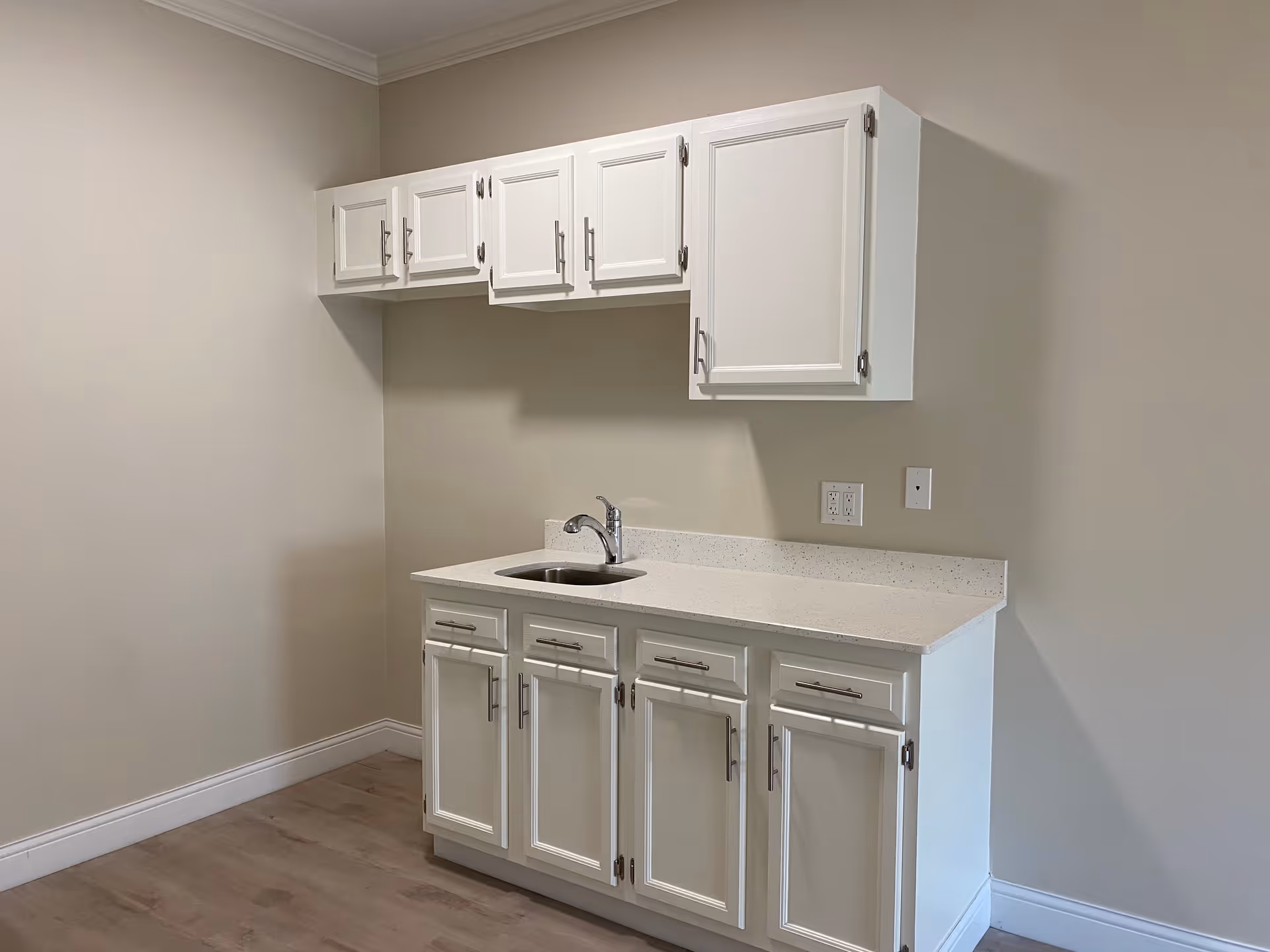 A small kitchenette area with white cabinets above and below a white countertop. There is a small sink with a faucet in the countertop. The walls are painted beige and the floor has light wood flooring.