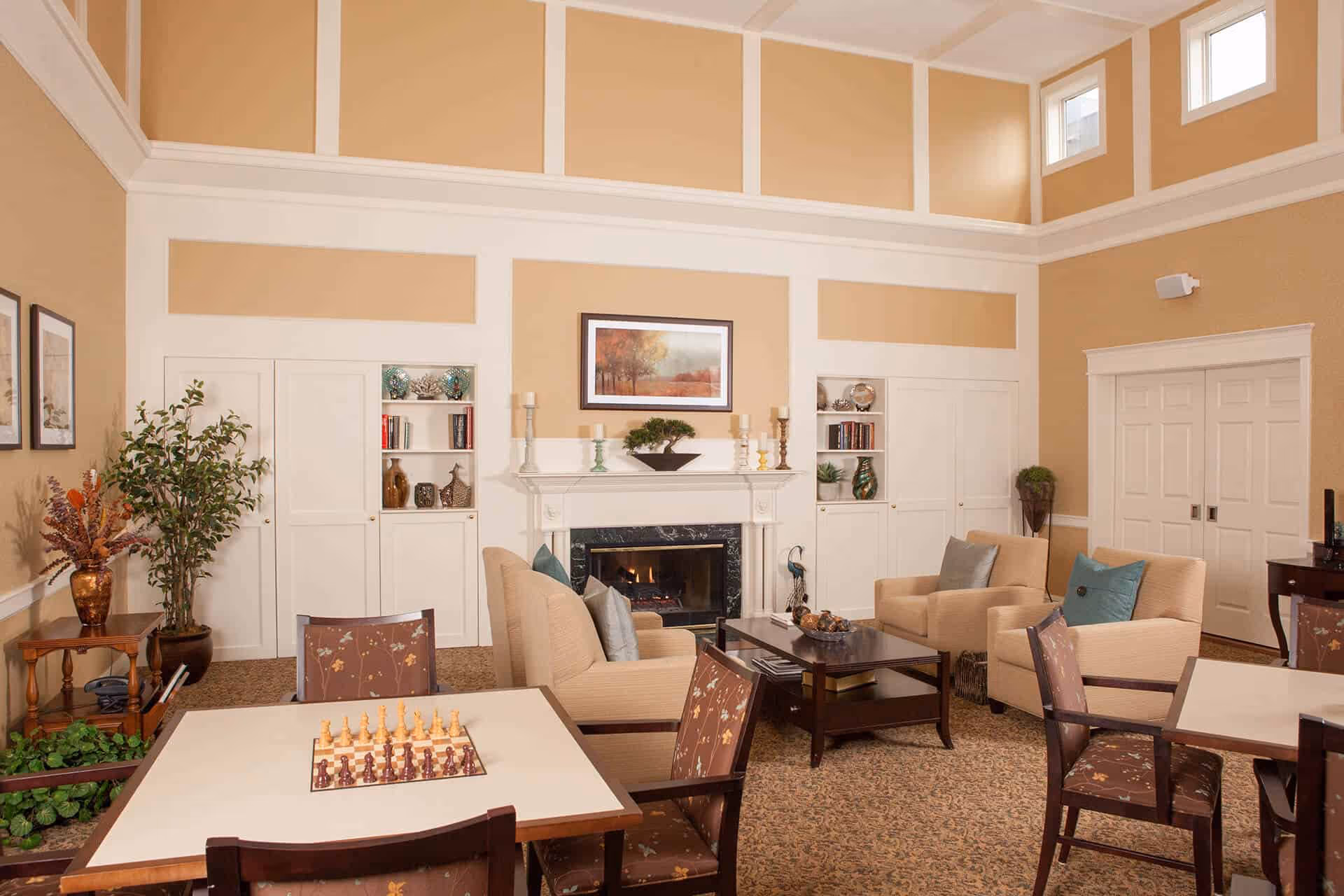 Spacious common living room with beige armchairs, coffee tables, a fireplace, built-in shelving, and a chessboard on a table.