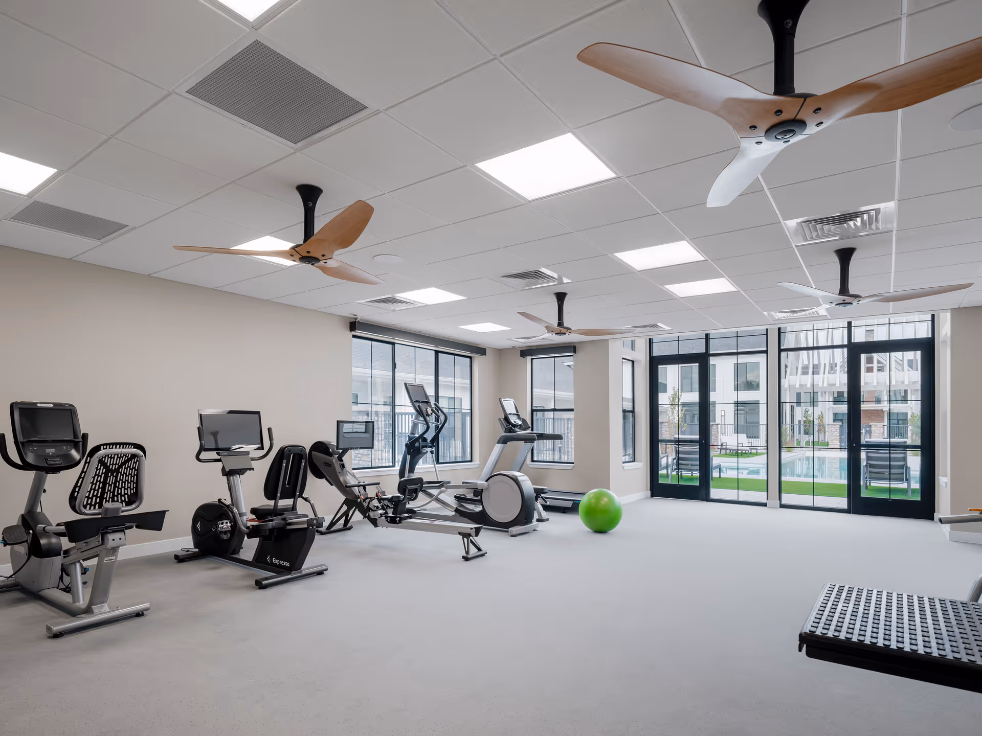 A bright and spacious fitness room with exercise equipment including stationary bikes, a rowing machine, and an elliptical trainer. The room has large windows and glass doors that provide a view of an outdoor area with a pool. The ceiling features modern wooden ceiling fans and recessed lighting.