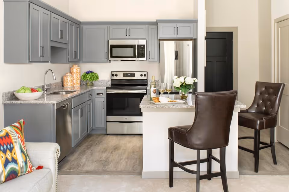 Modern kitchen with gray cabinets, stainless steel appliances including a microwave, stove, dishwasher, and refrigerator. A granite countertop island with two brown leather bar stools, a vase with white flowers, a bottle of wine, two wine glasses, and a plate with snacks. Part of a light-colored sofa with a colorful patterned pillow is visible on the left.