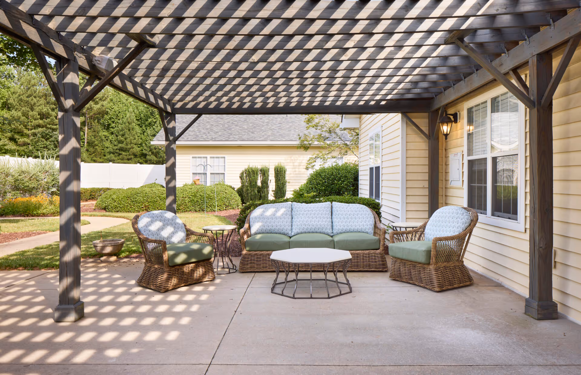 Outdoor patio area with a wooden pergola casting shadows on the concrete floor. There is a wicker sofa with green cushions and patterned back cushions, flanked by two matching wicker armchairs with green cushions. A small round side table and a hexagonal coffee table with metal legs are placed in front of the seating. The patio is adjacent to a yellow building with white-framed windows, and there are green bushes and trees in the background.