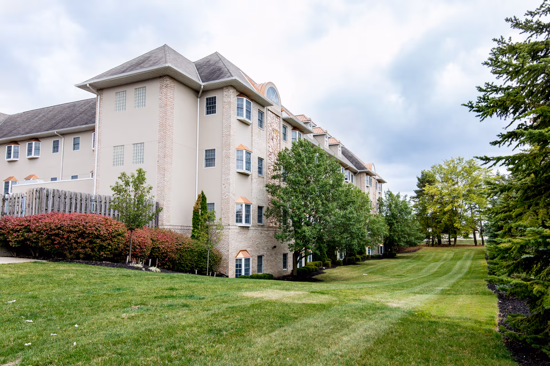 Exterior view of a multi-story senior living facility building with beige walls, multiple windows, and a well-maintained lawn with trees and shrubs under a cloudy sky.