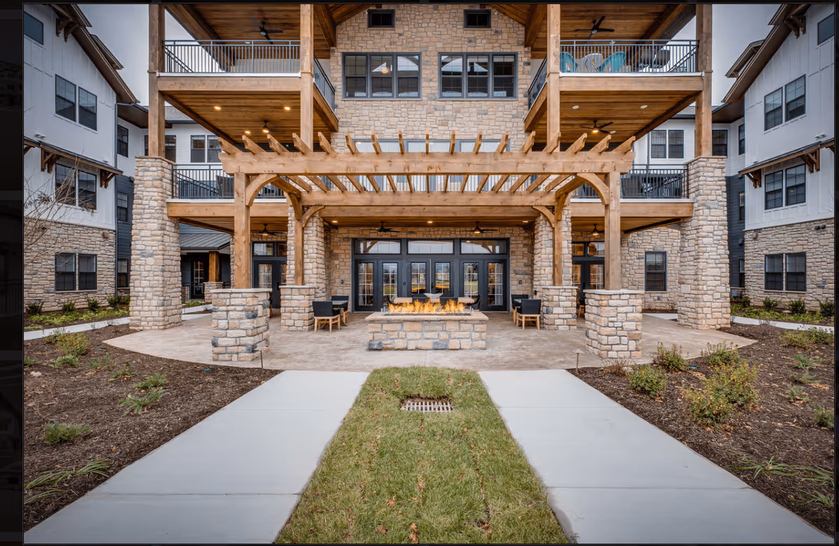Outdoor patio area of a senior living facility featuring a stone fire pit with flames, surrounded by wooden pergola and stone pillars. The building has multiple balconies with seating and large windows, and there are pathways and landscaped garden beds leading to the patio.