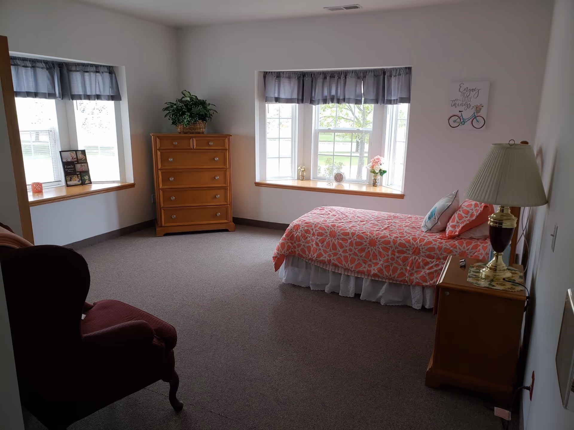 A cozy bedroom with a single bed covered in a coral and white patterned bedspread, two pillows, a wooden nightstand with a lamp, a wooden chest of drawers with a plant on top, two large windows with gray valances, and a burgundy armchair in the corner.