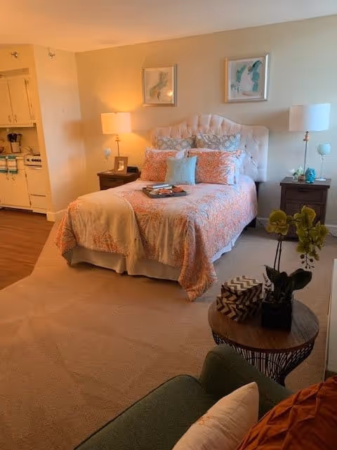 A cozy bedroom with a neatly made bed featuring a tufted headboard, coral and white bedding, and decorative pillows. Two nightstands with lamps flank the bed. There are two framed artworks above the headboard. In the foreground, a round wooden table holds a potted orchid and decorative items. Part of a green armchair with a pillow is visible. The room has beige carpet and a partial view of a kitchen area with white cabinets and appliances.