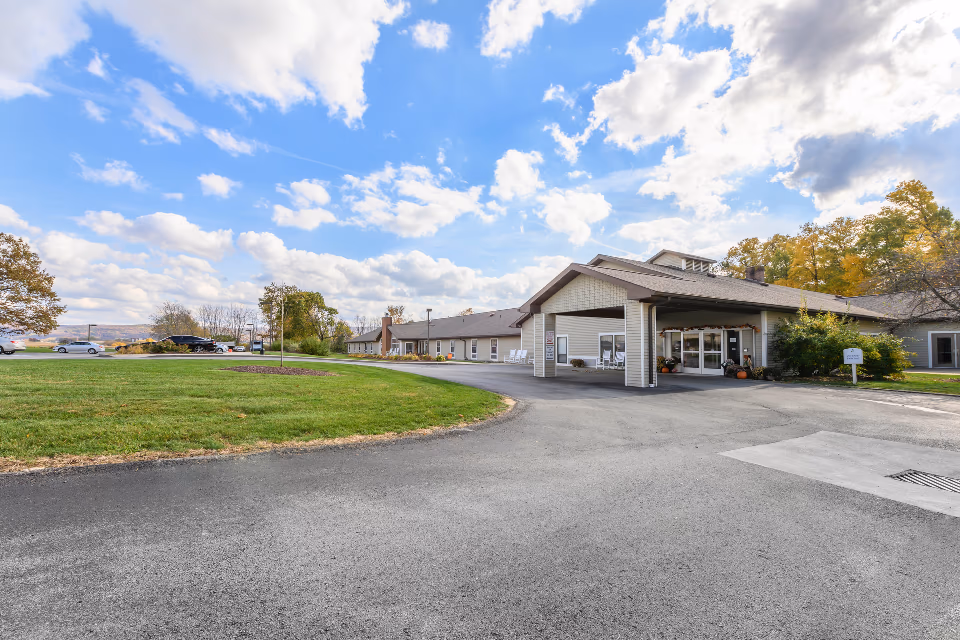 Exterior view of a single-story senior living facility building with a covered entrance, surrounded by a paved driveway and green lawn under a partly cloudy blue sky.