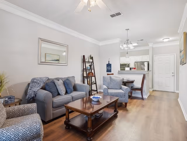 A cozy living room with a gray sofa, two patterned armchairs, a wooden coffee table with a decorative bowl, and a ladder-style shelf against the wall. The room has light-colored walls, a ceiling fan, and hardwood flooring. In the background, there is a small dining area with a round table and chairs, and a kitchen with white cabinets and stainless steel appliances.