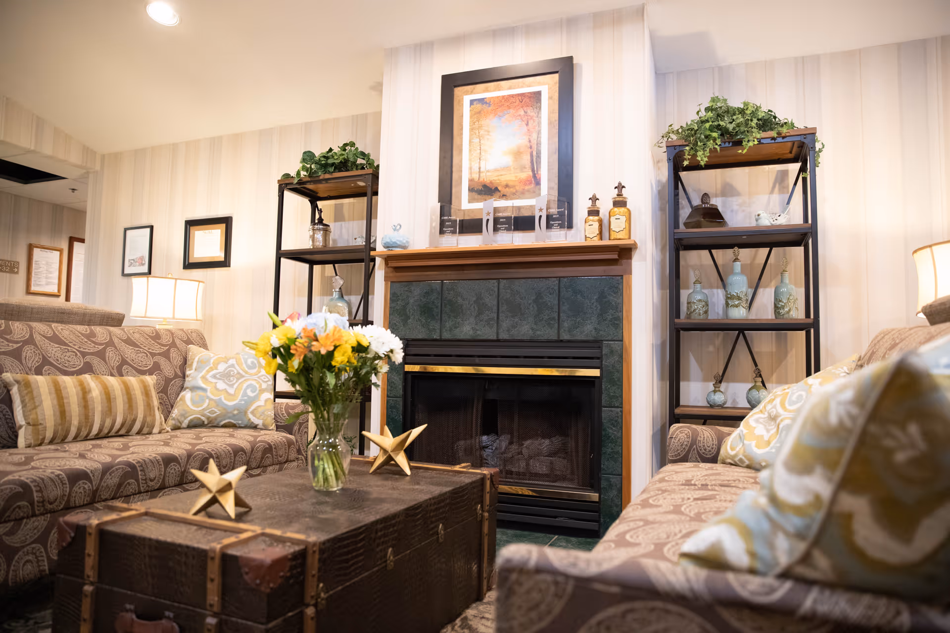 A cozy living room area with two patterned sofas facing each other, a dark wooden trunk used as a coffee table with a vase of fresh flowers and decorative stars on top, a fireplace with green tiles and a wooden mantel, two black metal shelves with decorative items and plants, and framed artwork and pictures on the walls.