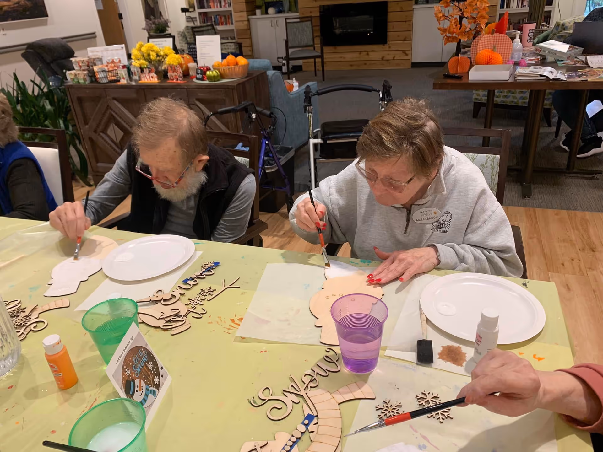 Two elderly individuals seated at a table engaged in painting wooden craft shapes. The table is covered with a light green tablecloth and has various craft supplies including paintbrushes, paint bottles, wooden cutouts, and plastic cups with water. The background shows a cozy interior with furniture, plants, and decorations.