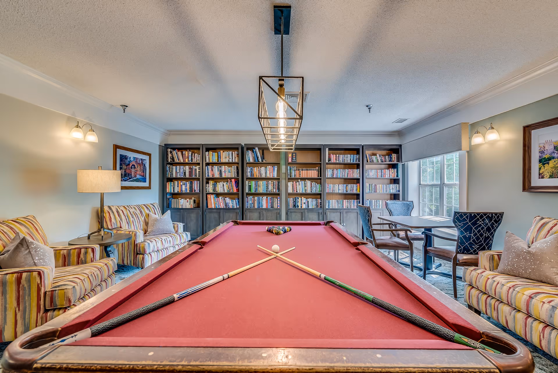 A cozy recreational room featuring a red pool table with two crossed cues and a white cue ball in the center. Behind the pool table is a large bookshelf filled with books. The room has comfortable striped armchairs and sofas on both sides, a table with four chairs near a window, and warm lighting from wall sconces and a ceiling light fixture.