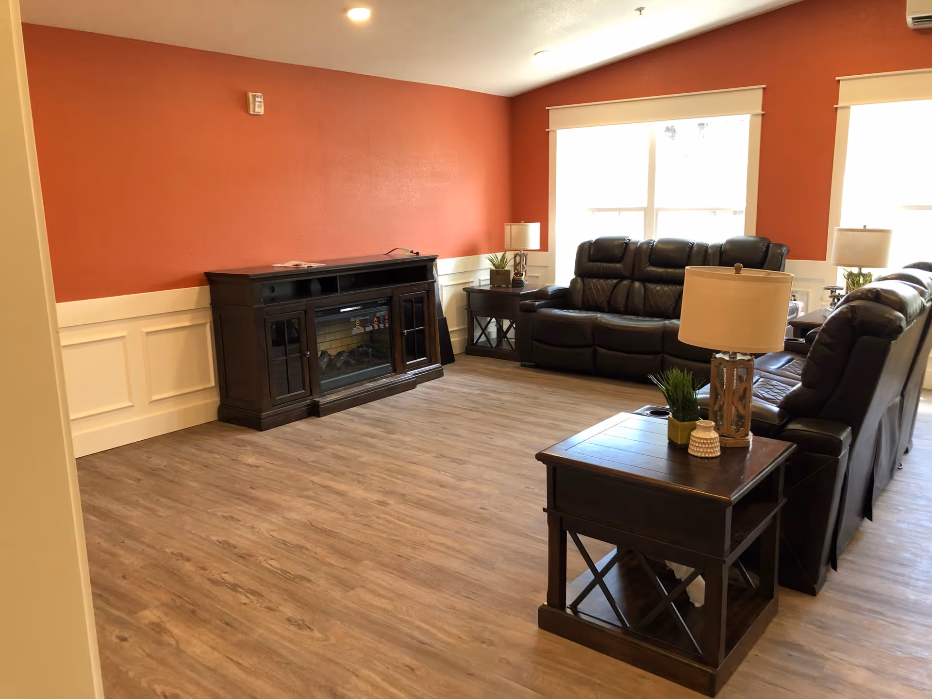 A living room with wood flooring, a dark wood electric fireplace against a red and white paneled wall, two dark leather sofas, two side tables with lamps, and two large windows letting in natural light.