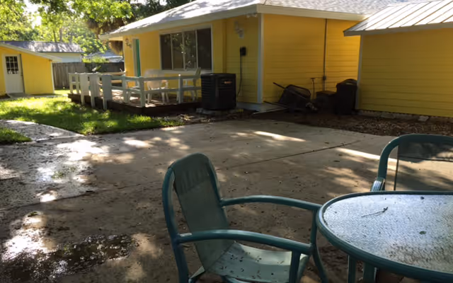 Outdoor patio area with a round glass table and green metal chairs in the foreground. In the background, there is a yellow building with a white railing and a small porch, surrounded by trees and grass.