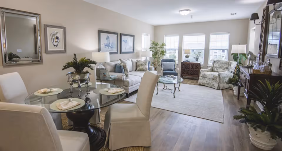A bright and spacious living room with a glass-top dining table set for four on the left, a beige sofa with decorative pillows, two armchairs, a glass coffee table, and several lamps and plants. The room has large windows letting in natural light and wooden flooring.