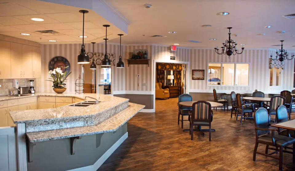 Interior view of a senior living facility dining area with multiple tables and chairs, a kitchen counter with a sink and hanging pendant lights, striped wallpaper, wooden flooring, and chandeliers. A cozy sitting area with armchairs is visible through an open doorway.