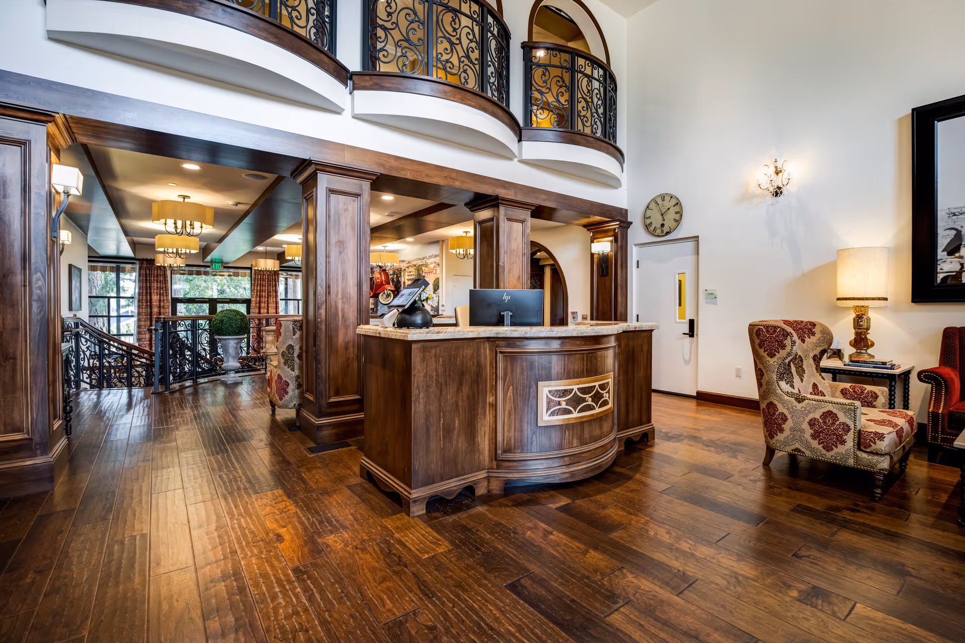 Interior view of a senior living facility reception area with a wooden reception desk, ornate iron railings on the upper balcony, hardwood floors, upholstered armchairs, a wall clock, and warm lighting fixtures.