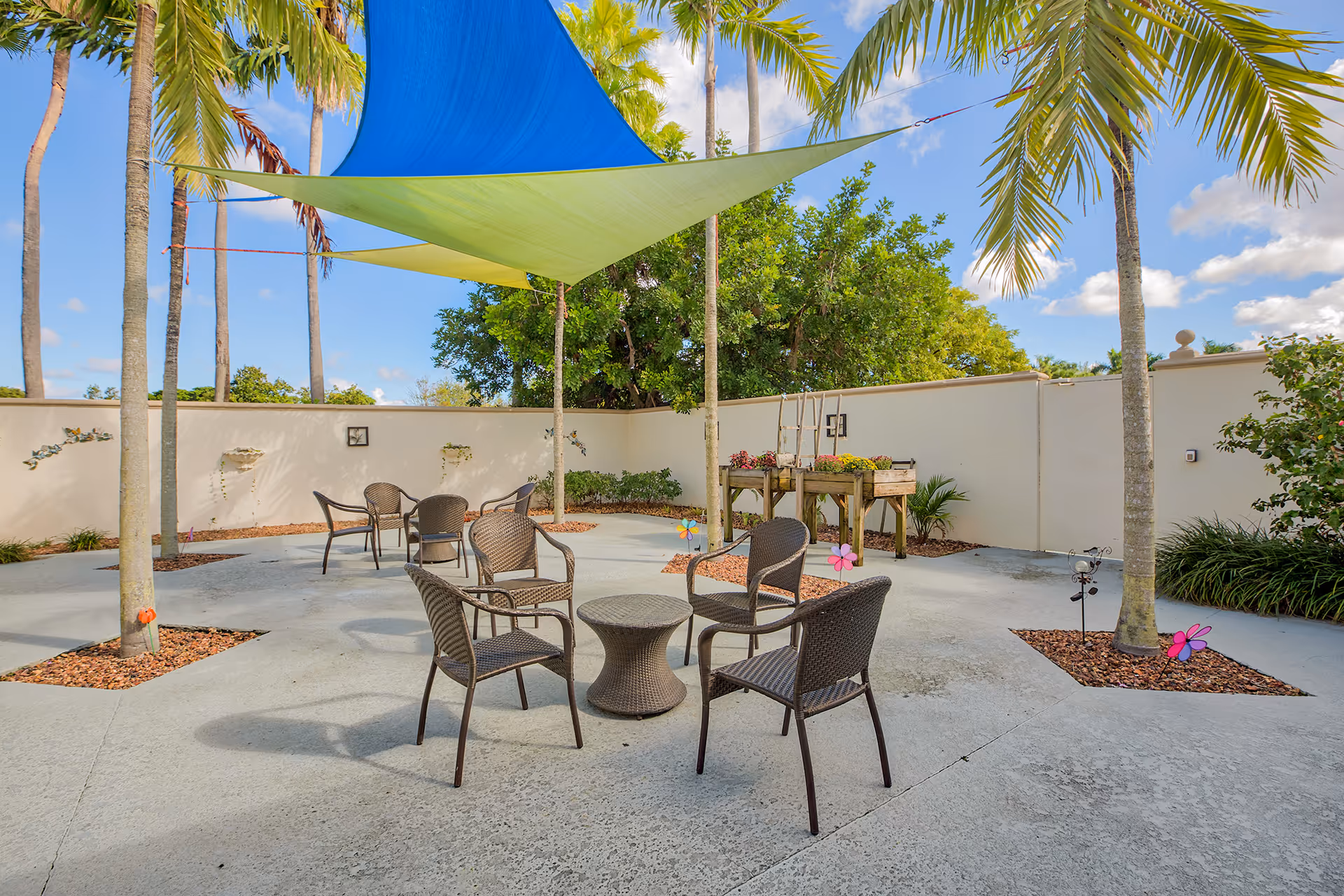 Outdoor patio area with several brown wicker chairs arranged around small tables on a concrete surface. There are palm trees planted in square cutouts in the concrete, and colorful triangular shade sails in blue and green are stretched overhead. The area is enclosed by a beige wall with some greenery and decorative elements along the edges.