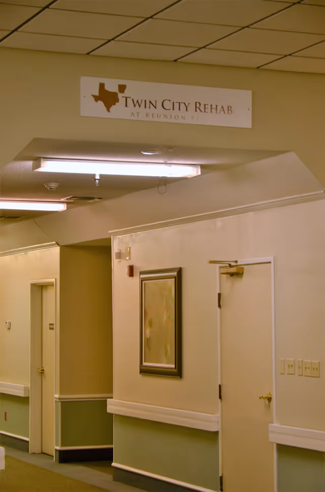 Interior hallway of a senior care facility with beige and green walls, a framed abstract painting, two closed doors, and a ceiling sign that reads 'Twin City Rehab at Reunion Plaza'.