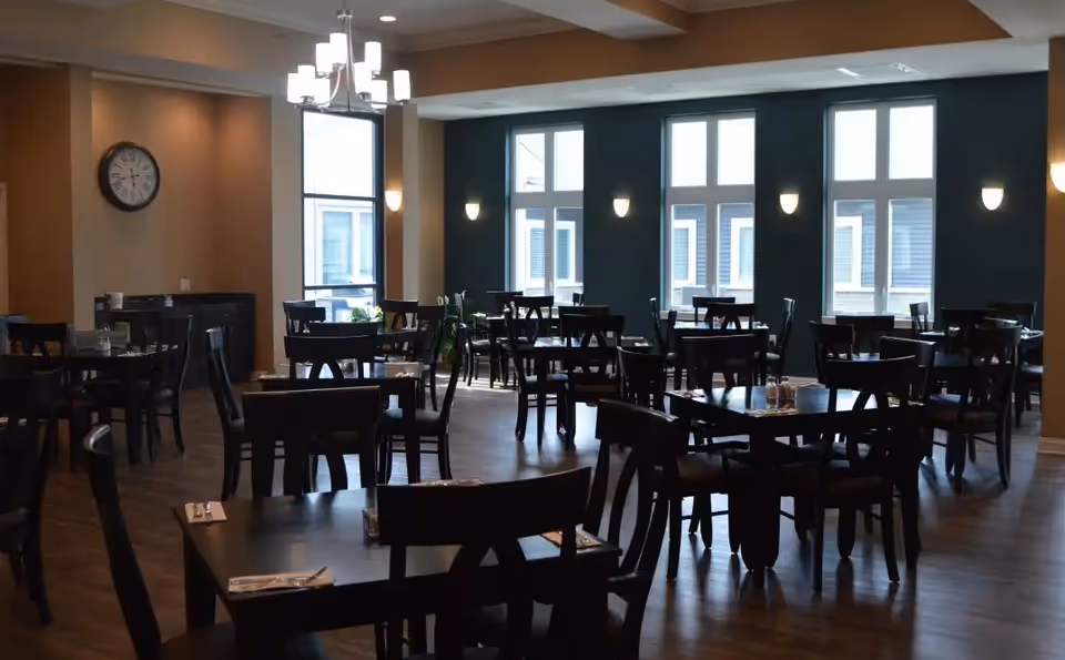 Dining room with dark wood tables and chairs arranged under chandeliers and large windows.