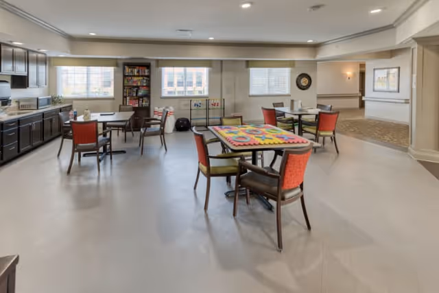 A spacious, well-lit common area with several tables and chairs arranged for group activities or dining. One table has a colorful puzzle on it. The room features large windows letting in natural light, a bookshelf filled with games and books, and a kitchenette area with dark cabinets and a countertop along one wall.
