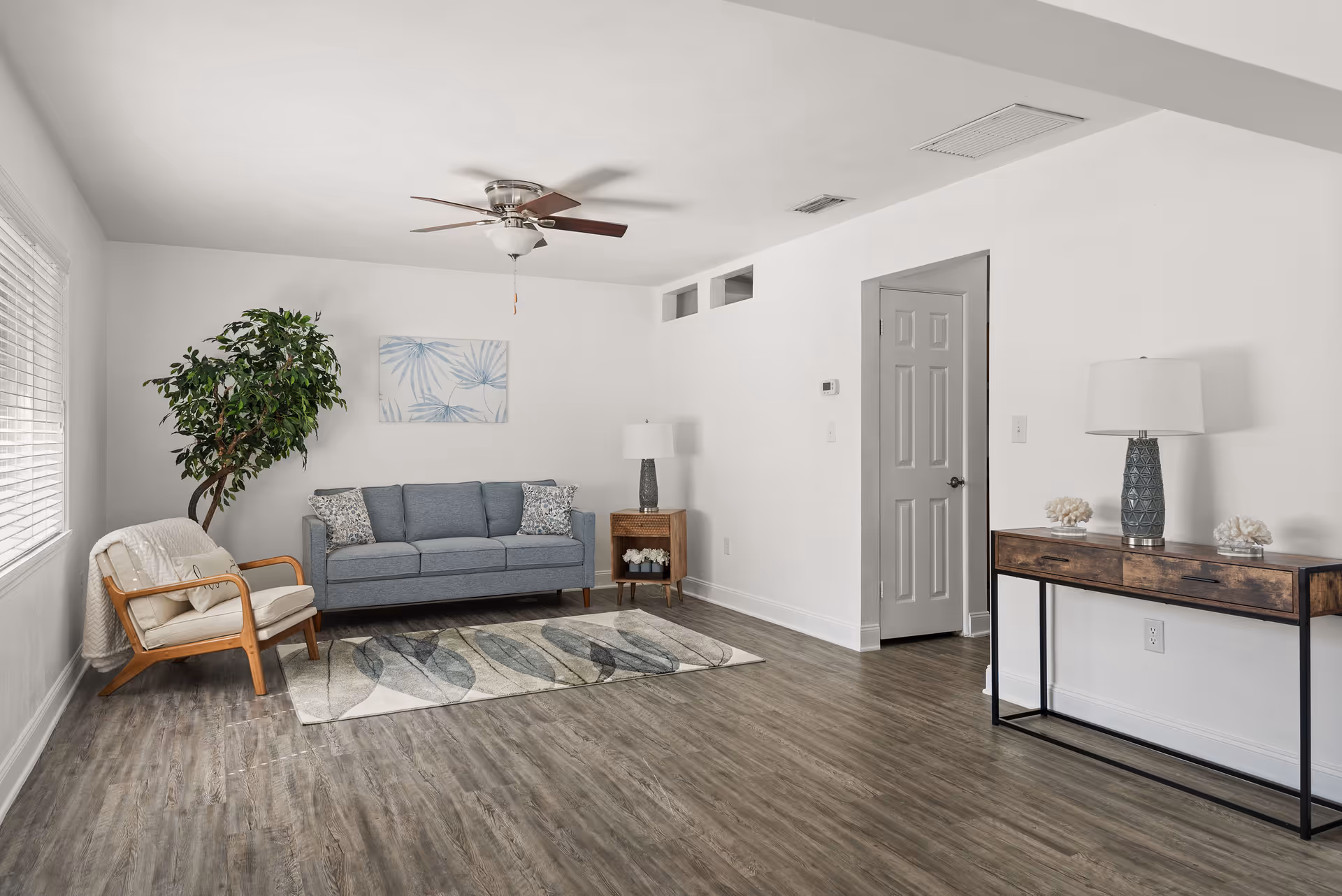 Bright minimalist living room with a blue sofa, wooden armchair, rug, console table, potted plant, and ceiling fan.
