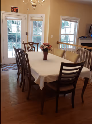 A dining area with a rectangular table covered with a white tablecloth and surrounded by eight chairs. A vase with flowers is placed in the center of the table. The room has wooden flooring, two large windows, and a glass door with an exit sign above it. There is a railing separating the dining area from a living space with a fireplace and television.