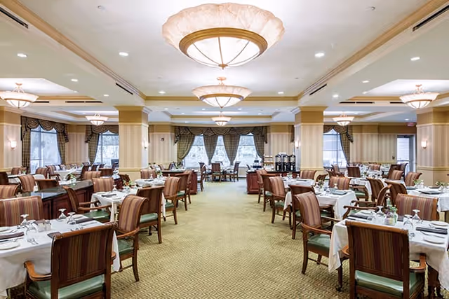 A spacious and elegant dining room with multiple tables covered in white tablecloths, each set with plates, glasses, and silverware. The room features large windows with draped curtains, several ceiling light fixtures, and comfortable wooden chairs with striped upholstery.