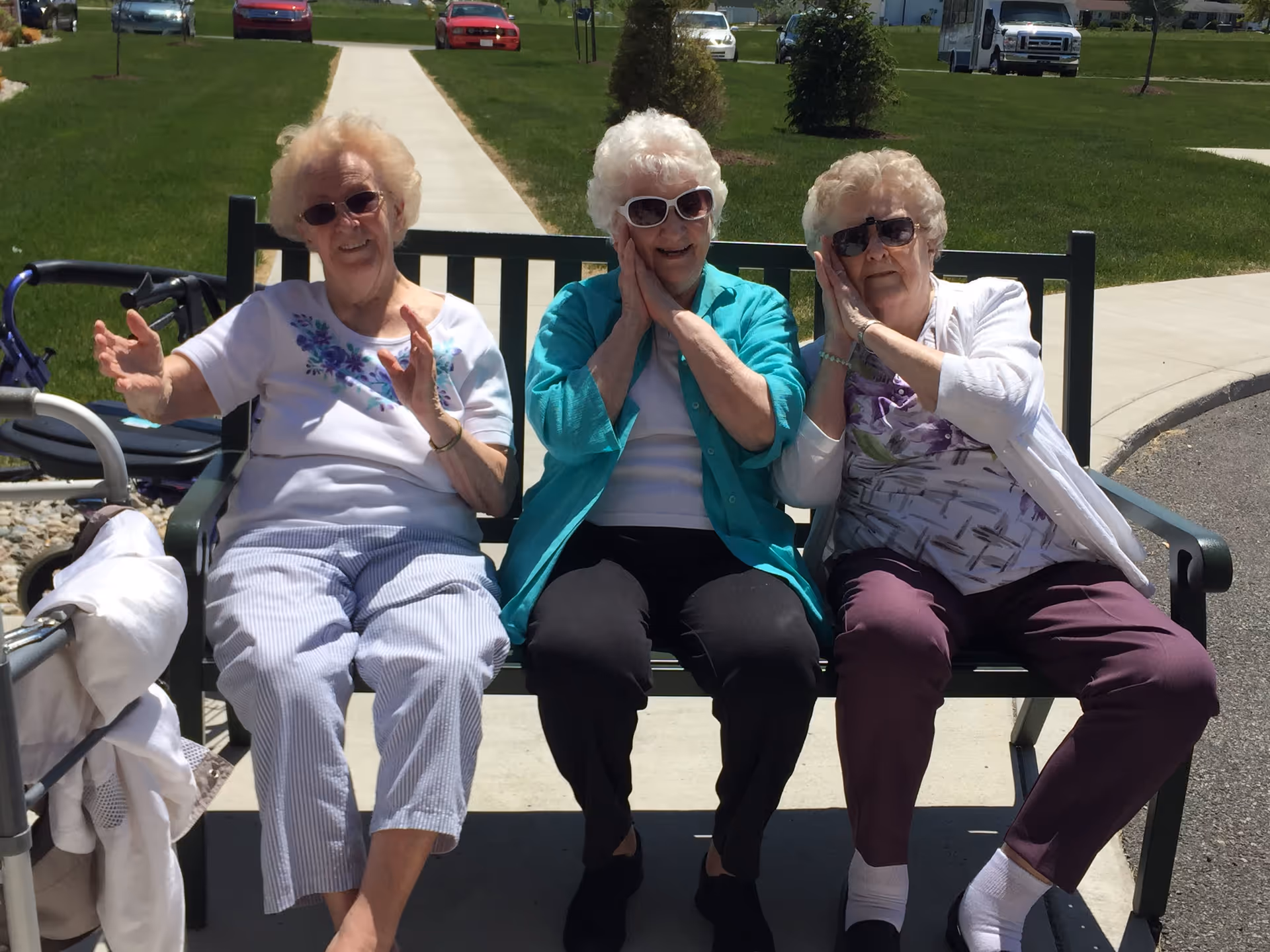 Three elderly women sitting on a bench outdoors on a sunny day, each wearing sunglasses and making playful hand gestures near their faces. Behind them is a sidewalk, green grass, and parked cars.