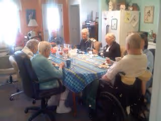 A group of elderly people sitting around a rectangular dining table covered with a blue checkered tablecloth, enjoying a meal together in a well-lit room with a refrigerator and kitchen items in the background.