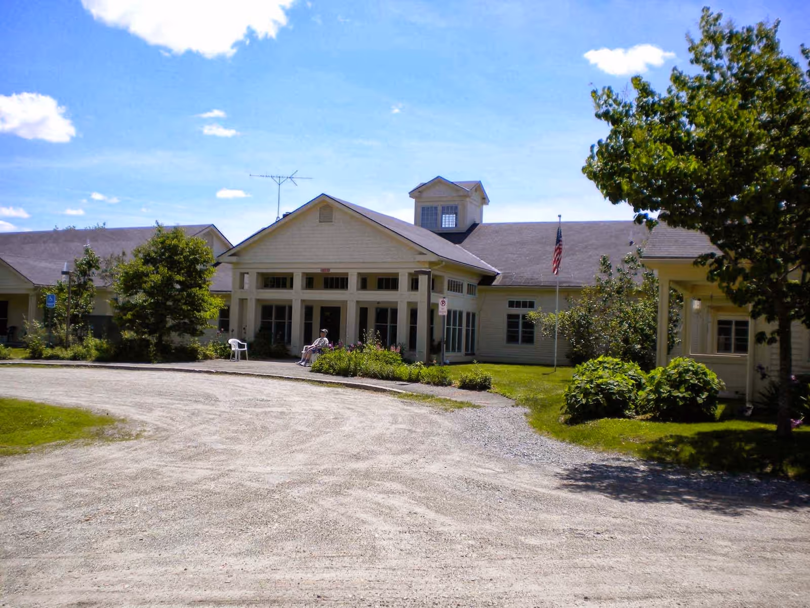 Front entrance of a single-story senior care building with a gravel driveway, landscaping, and an American flag.