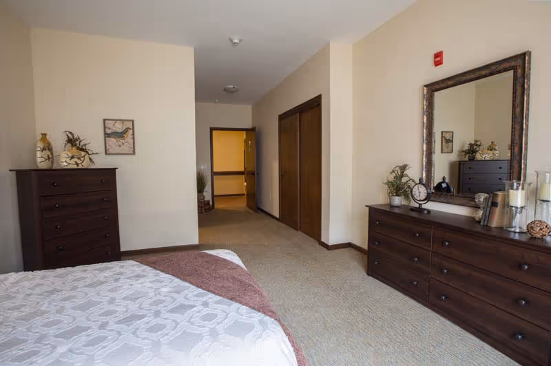 A bedroom in a senior living facility with a bed covered in a white and beige patterned bedspread and a brown throw blanket. The room features two dark wooden dressers, one with a large mirror above it, decorated with plants, candles, and a clock. The walls are light beige, and there is a framed bird picture on the wall. The room has a carpeted floor and a hallway with wooden closet doors leading to another area.