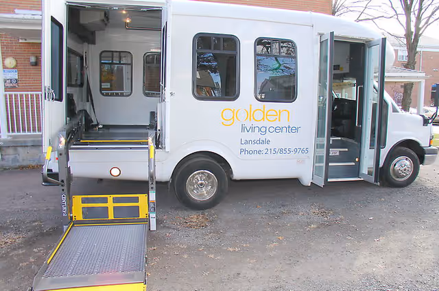 A white shuttle bus with an extended wheelchair lift parked outside a building. The bus has the text 'golden living center Lansdale Phone: 215/855-9765' on its side. The bus doors are open, showing the interior seating area.