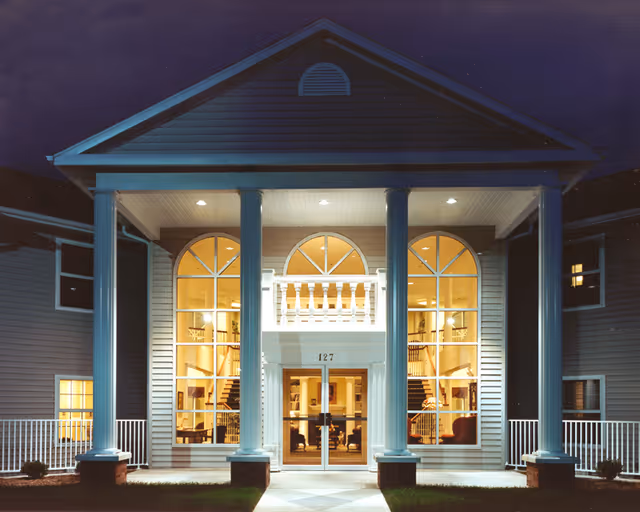 Night view of the front entrance of a two-story building with large white columns, arched windows, and warm interior lighting. The building has a pitched roof and the number 127 above the glass double doors.