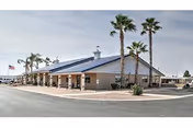 Exterior view of a single-story building with a sloped roof and several palm trees in front. The building has a covered walkway supported by columns and an American flag flying on a flagpole to the left. The sky is clear and the surrounding area is paved.